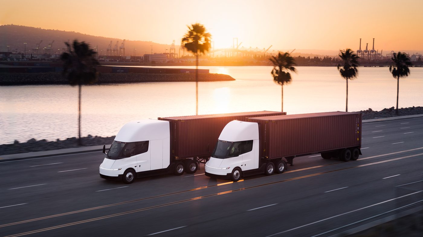 Two Tesla Semi trucks hauling containers along a coastal California highway at sunset, with palm trees and port cranes in the background