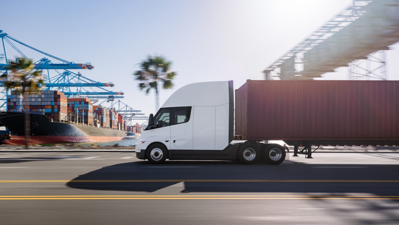Tesla Semi hauling a container past shipping cranes and a cargo vessel at the port, palm trees along the road
